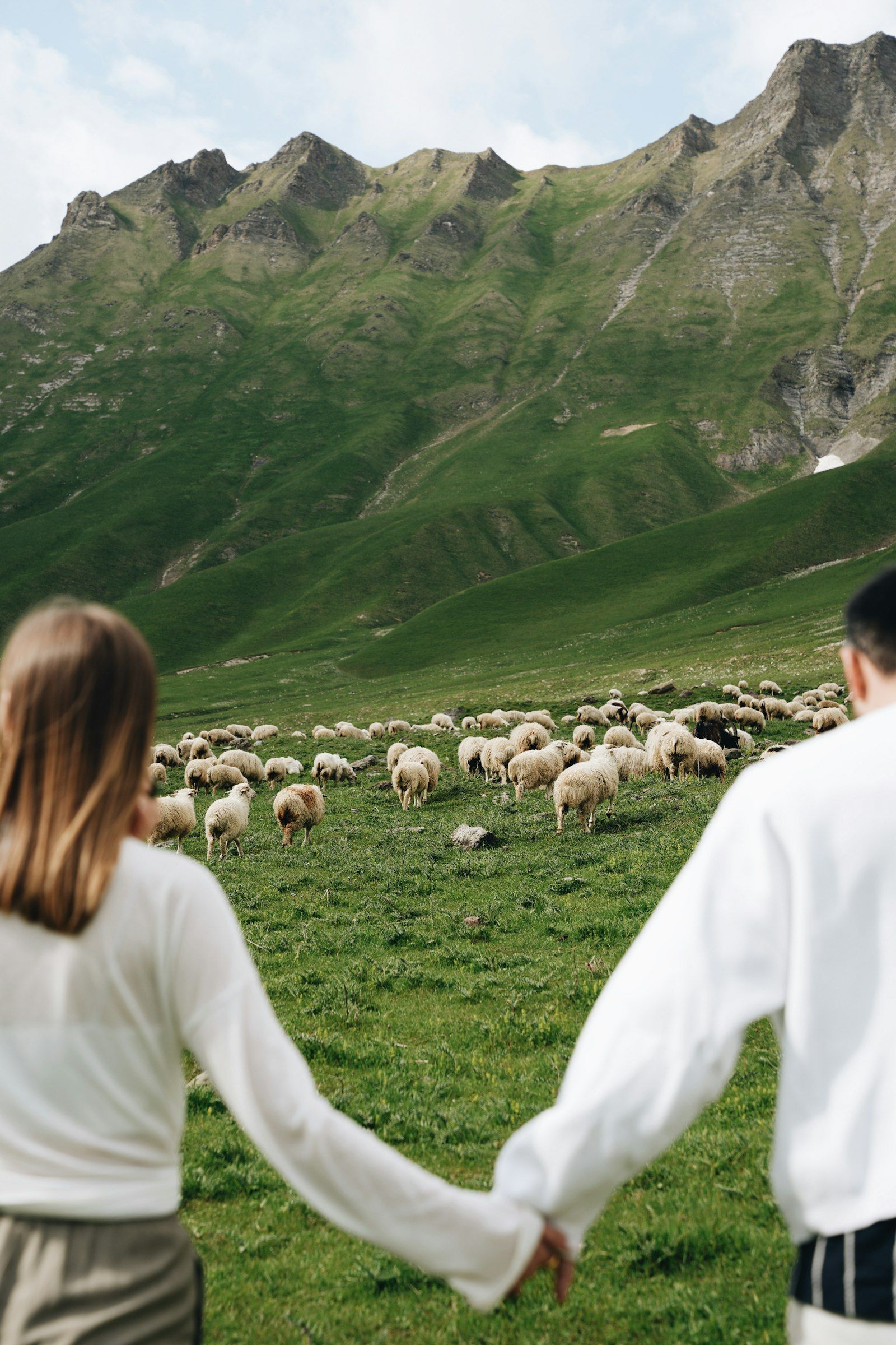A couple holding hands while a shepherd’s flock grazes beneath the peaks above Gudauri